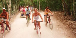 Mulher andando de bicicleta por ruas de terra em Cobá, cercada por vegetação tropical.