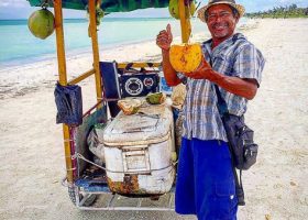 Homem numa barraca a vender cocos em Holbox, com ambiente tropical e praia ao fundo.